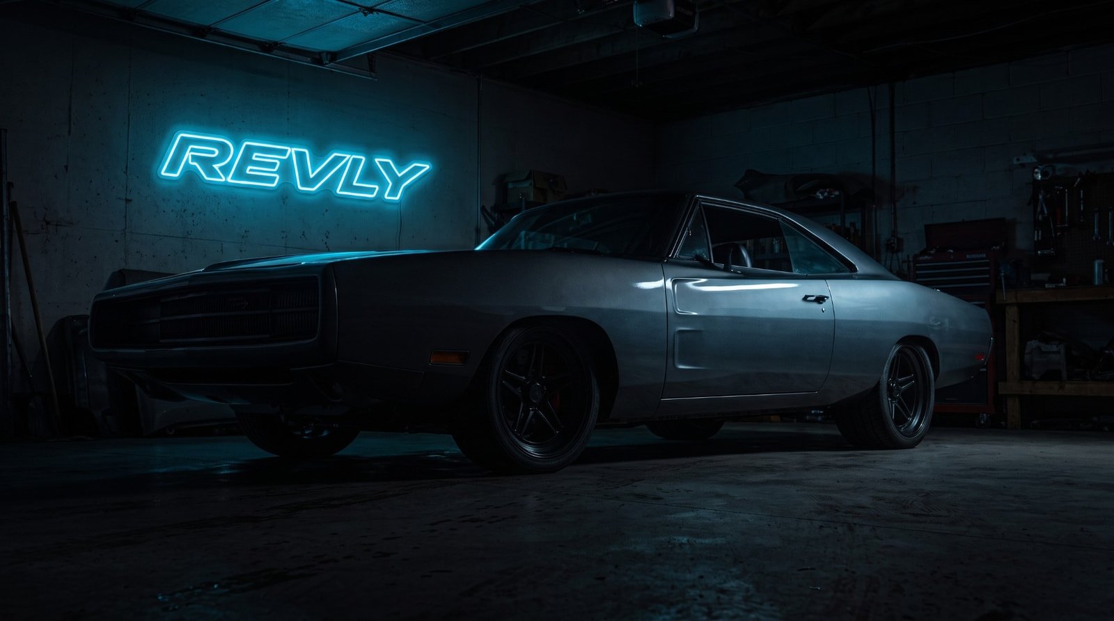 A gray classic muscle car in a dark garage, lit by a glowing blue REVLY neon sign